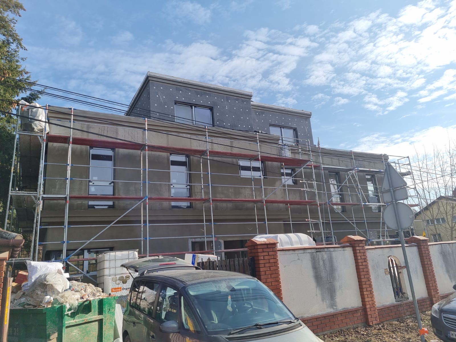 Budynek w trakcie ocieplania, otoczony rusztowaniami. Widoczne płyty izolacyjne na górnej części elewacji, śmietnik z odpadami budowlanymi i zaparkowane samochody.