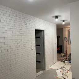 Ściana w korytarzu po położeniu tapety imitującej białą cegłę, widoczna szara stolarka drzwiowa, czarne lampy sufitowe i piec w tle, podłoga zabezpieczona kartonem, widać narzędzia malarskie.