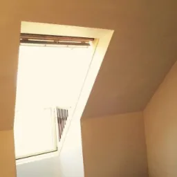 Poddasze po szpachlowaniu i malowaniu, widoczne okno dachowe z żaluzjami, ślady gipsu na folii zabezpieczającej podłogę.