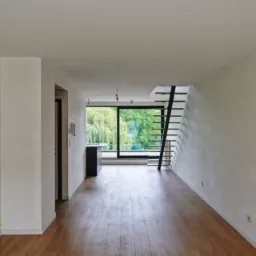 Wnętrze po remoncie: parkiet, świeżo pomalowane ściany w odcieniach bieli, nowoczesna klatka schodowa z czarnymi elementami, duże okno z widokiem na zieleń.