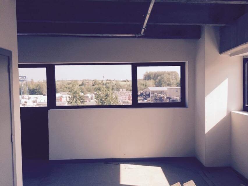 Pusty pokój z pomalowanymi na biało ścianami i oknami z widokiem na krajobraz miejski z zielenią, widoczne rury pod sufitem, brak mebli.