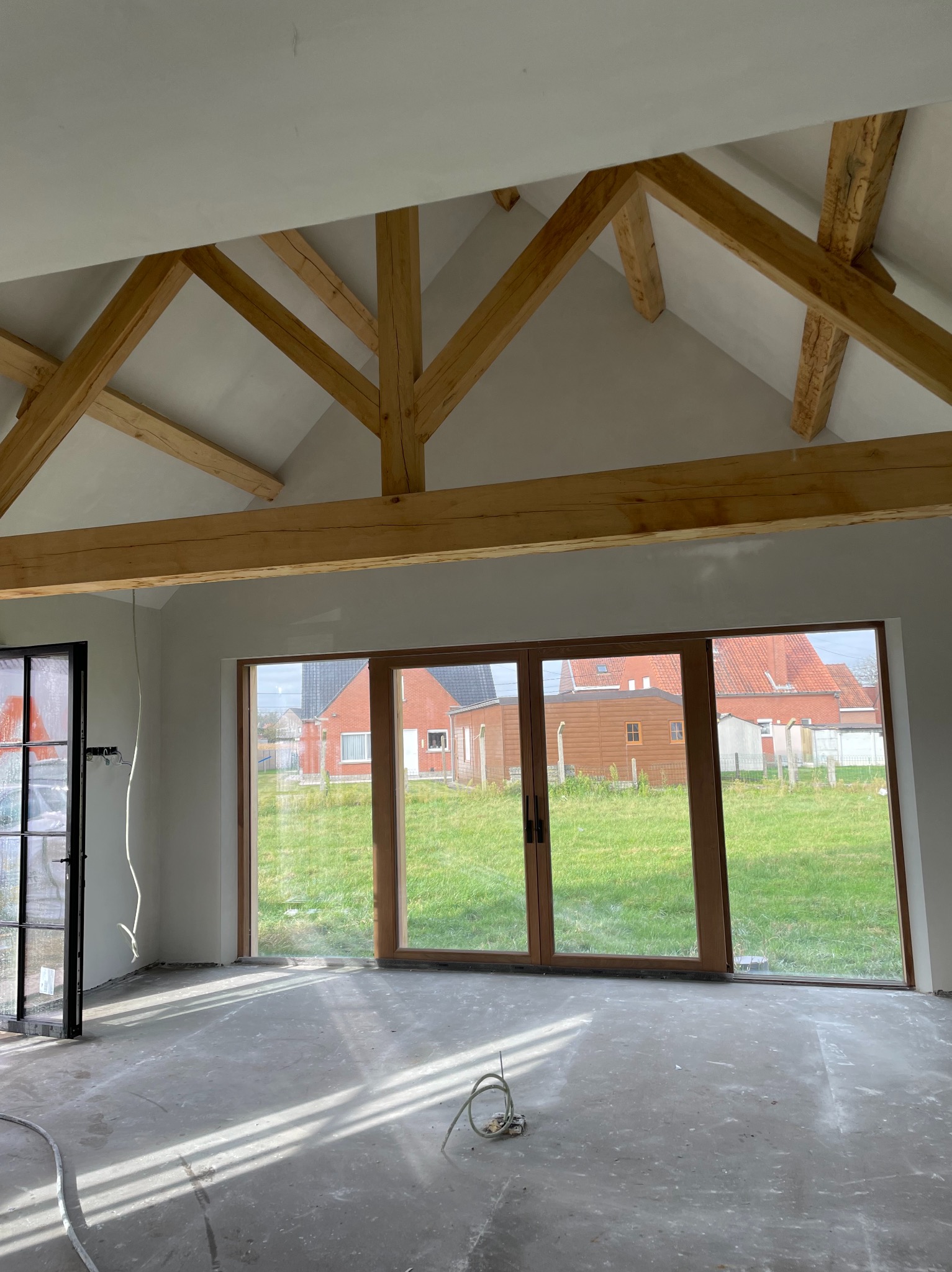 Wnętrze pokoju z drewnianą konstrukcją dachu, dużymi oknami przesuwnymi z widokiem na trawnik i budynki, oraz surową podłogą w trakcie prac wykończeniowych.