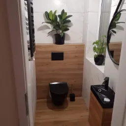 Nowoczesna łazienka w Rzeszowie po remoncie: czarna armatura, umywalka narożna z szafką z drewnopodobnego materiału, podwieszana toaleta, białe płytki imitujące marmur i drewniana zabudowa ściany...