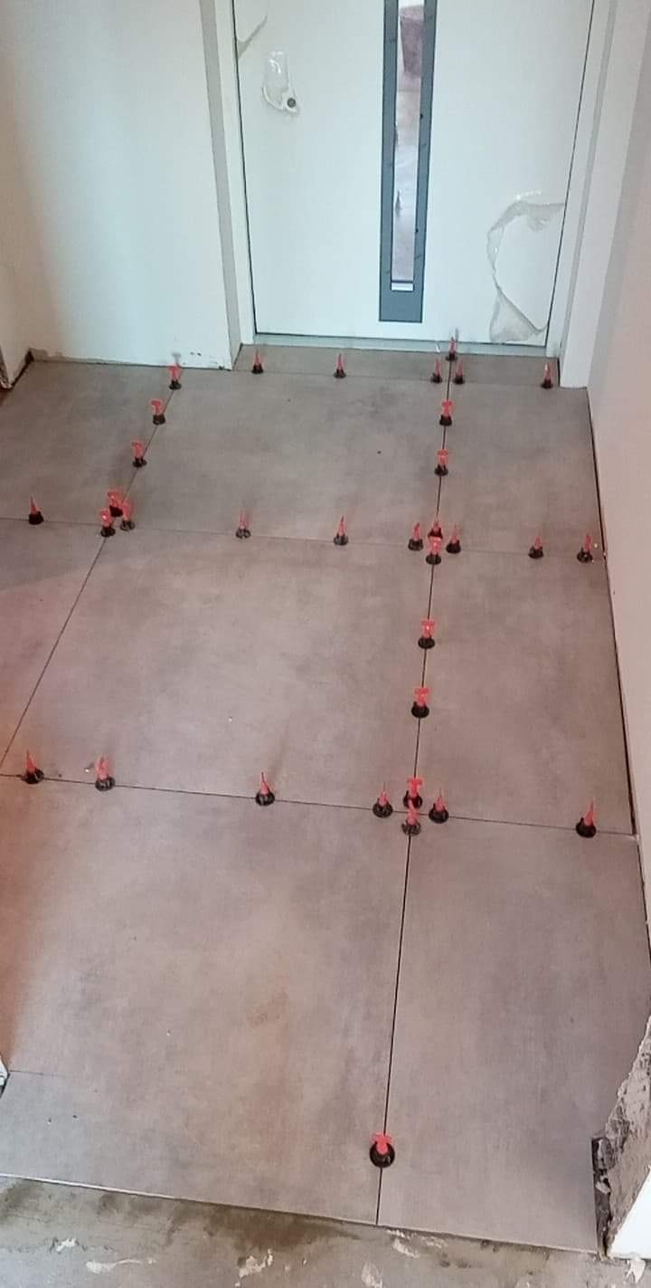 Układanie szarych płytek podłogowych w korytarzu, z użyciem systemu poziomowania z czerwonymi klinami i czarnymi podstawkami, widoczny fragment drzwi wejściowych.