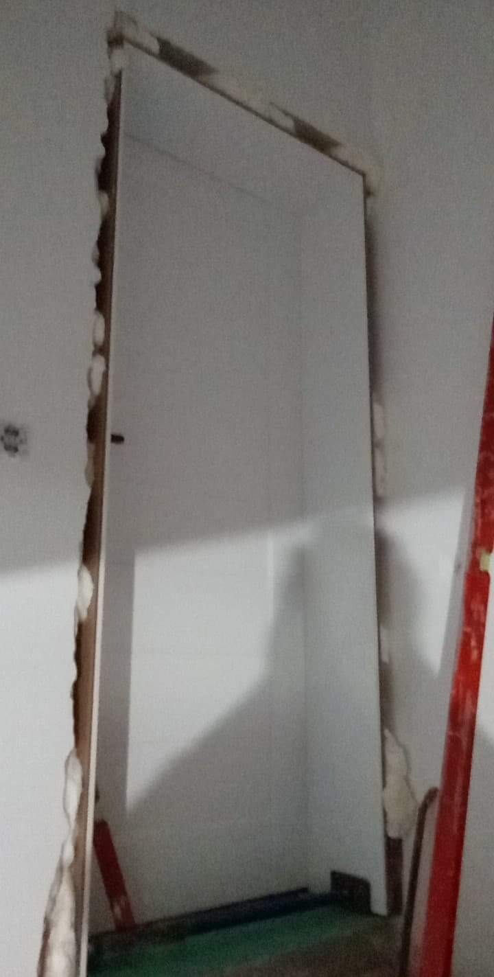 Niedokończony montaż ościeżnicy w surowym otworze ściany, widoczna pianka montażowa i fragment czerwonej poziomicy.