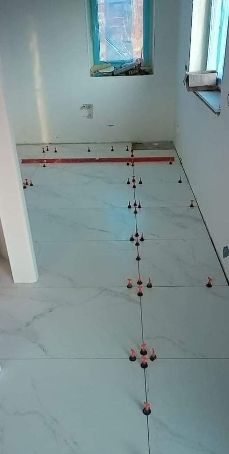 Układanie białych płytek podłogowych z marmurowym wzorem w łazience, z użyciem czerwonych klinów poziomujących i poziomicy. Okno zabezpieczone folią malarską.