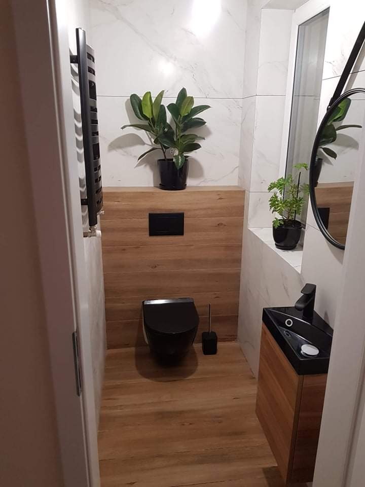 Nowoczesna łazienka w Rzeszowie po remoncie: czarna armatura, umywalka narożna z szafką z drewnopodobnego materiału, podwieszana toaleta, białe płytki imitujące marmur i drewniana zabudowa ściany...