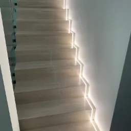 Drewniane schody z podświetleniem LED na każdym stopniu, zabezpieczone szklaną balustradą, widok z dołu.