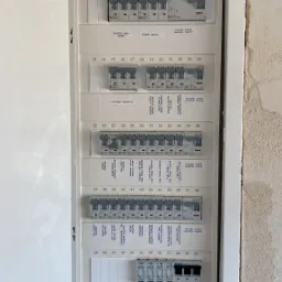 Otwarta skrzynka rozdzielcza z bezpiecznikami i opisami obwodów, widoczne wyłączniki różnicowoprądowe i ograniczniki przepięć, ściana w stanie surowym.