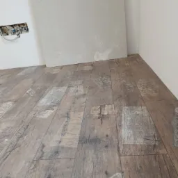 Ułożone panele podłogowe o wzorze imitującym stare drewno z nadrukami, widoczny fragment ściany z wystającą instalacją elektryczną i fragment płyty gipsowo-kartonowej.