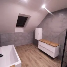 Wykończona łazienka na poddaszu ze skośnym sufitem, z szarymi płytkami na ścianach, drewnopodobną podłogą, białą wanną, grzejnikiem drabinkowym, szafką pod umywalkę z drewnianym blatem i kabiną...