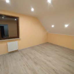 Poddasze po remoncie z jasnymi panelami podłogowymi imitującymi drewno, pomarańczowe ściany, biały grzejnik pod oknem z brązową ramą i wbudowane oświetlenie sufitowe.