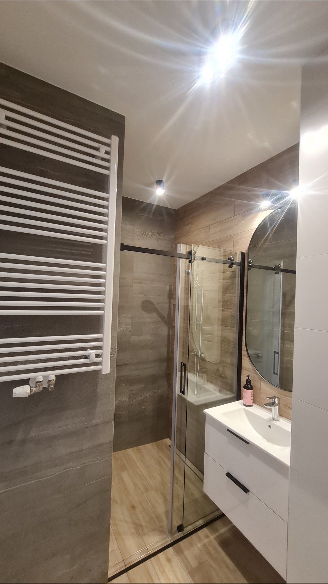 Nowoczesna łazienka z kabiną prysznicową typu walk-in z czarnymi profilami, białą szafką pod umywalkę z czarnymi uchwytami i grzejnikiem drabinkowym na ścianie z szarych płytek imitujących beton.