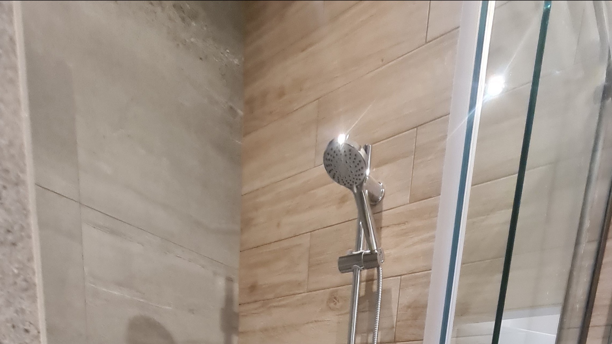 Chromowana słuchawka prysznicowa z regulacją strumienia, zamontowana na szynie w kabinie prysznicowej wykończonej płytkami imitującymi drewno, Gdańsk.