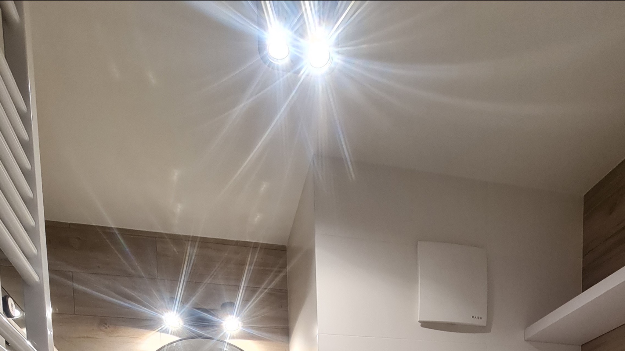 Jasne oświetlenie sufitowe w wykończonej łazience z elementami imitującymi drewno, widoczny biały grzejnik drabinkowy i wentylator ścienny.