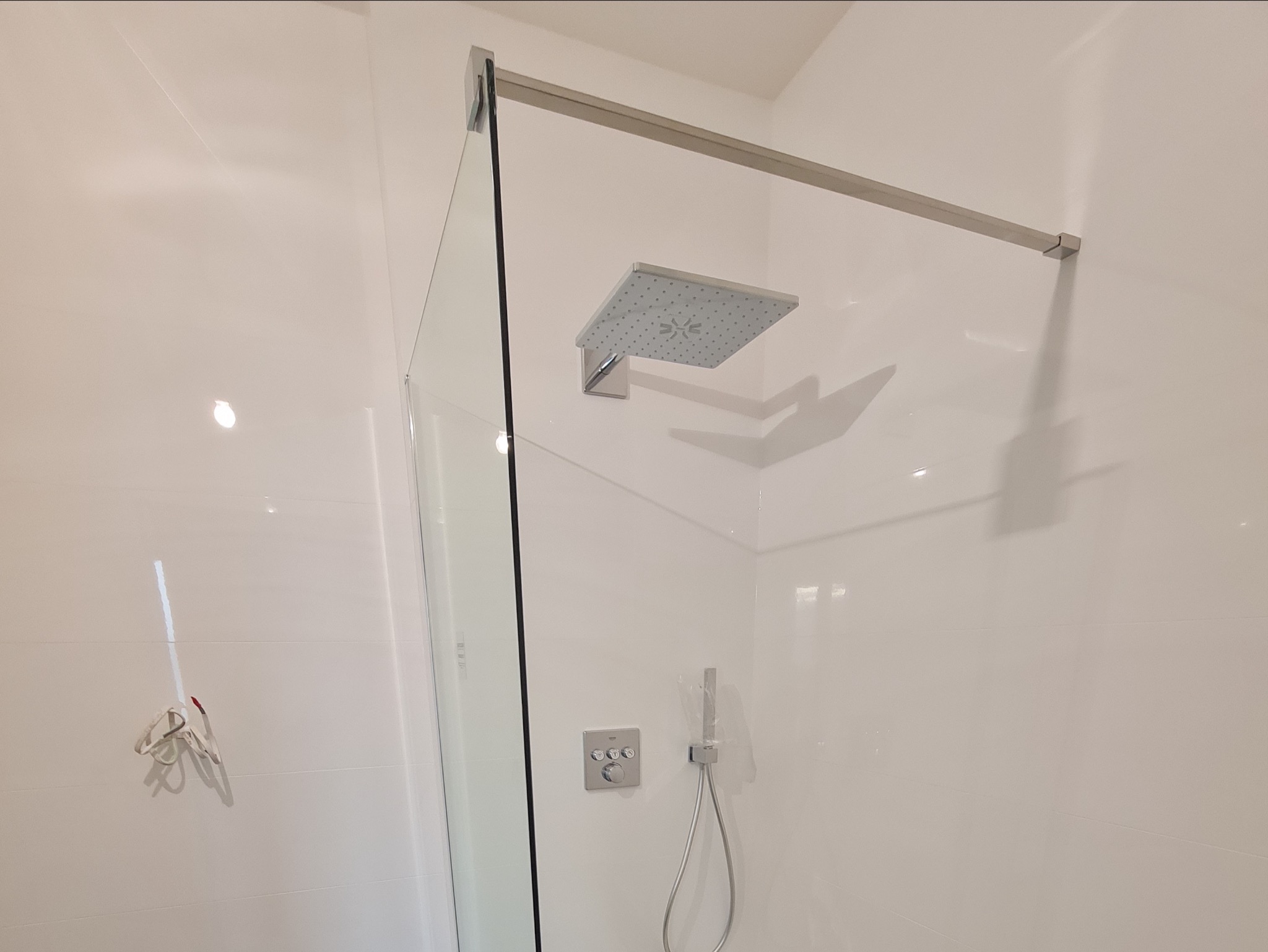 Nowoczesna kabina prysznicowa walk-in z kwadratową deszczownicą i minimalistyczną baterią podtynkową na białych, błyszczących płytkach.