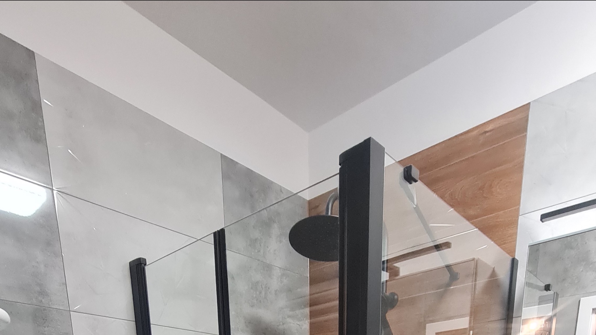 Kabina prysznicowa z czarnymi profilami i deszczownicą, widoczne płytki imitujące drewno i szare płytki w łazience.