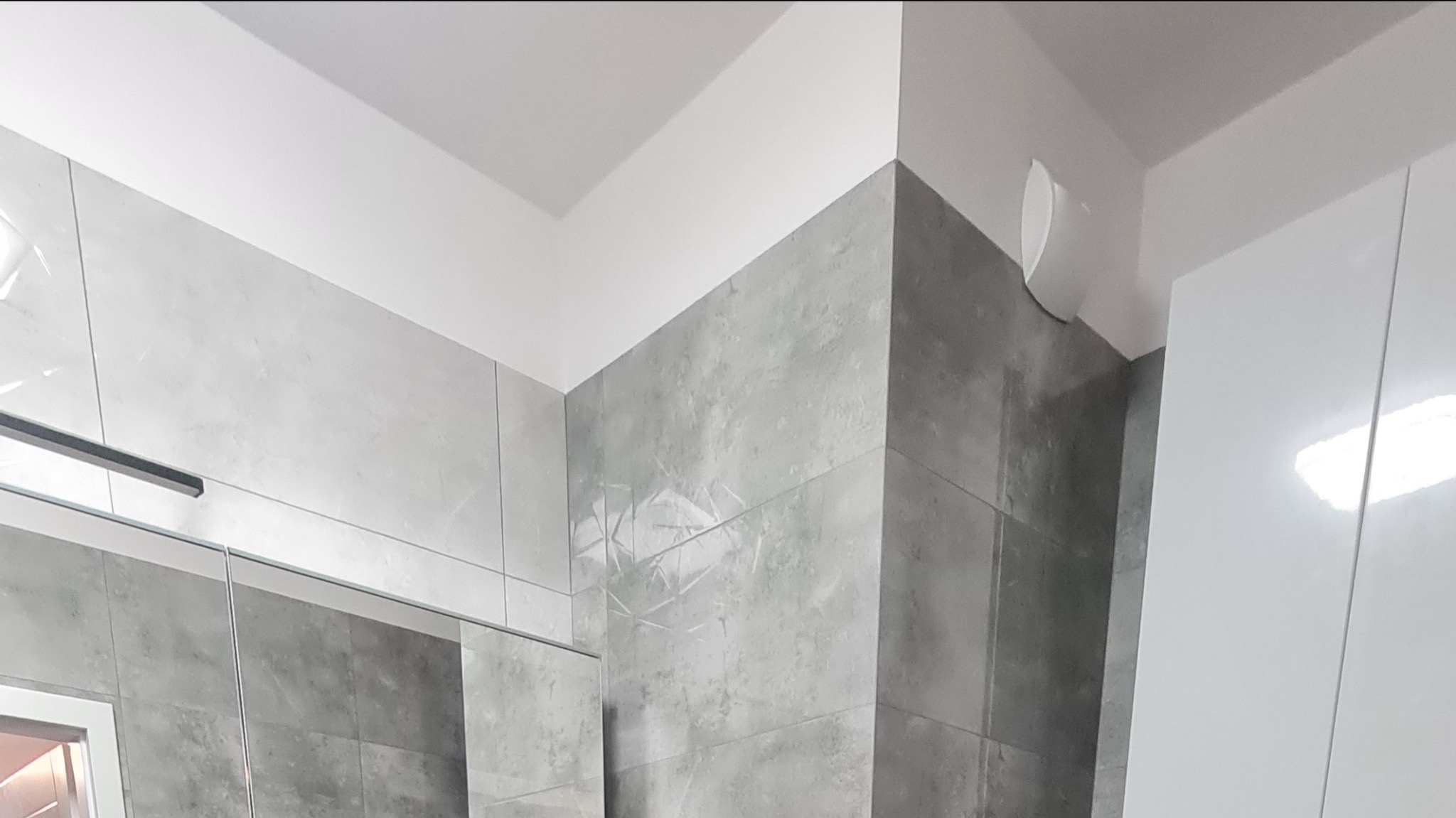 Fragment łazienki po remoncie z szarymi płytkami imitującymi beton, biały sufit i szafka, widoczny fragment lustra z czarną listwą oświetleniową.