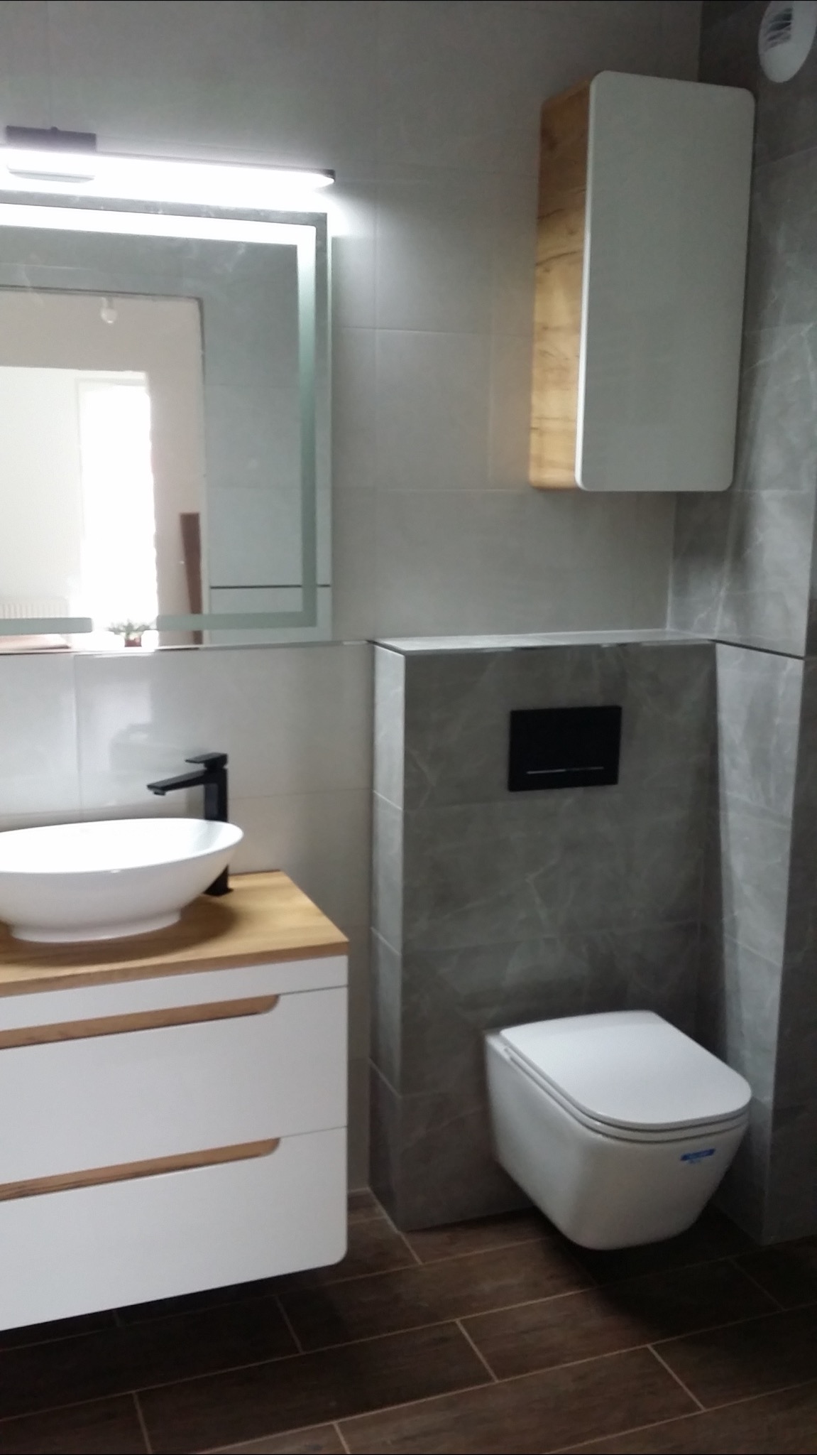 Nowoczesna łazienka w Gdańsku z umywalką nablatową na drewnianej szafce, podwieszaną toaletą i szarą płytką imitującą marmur.