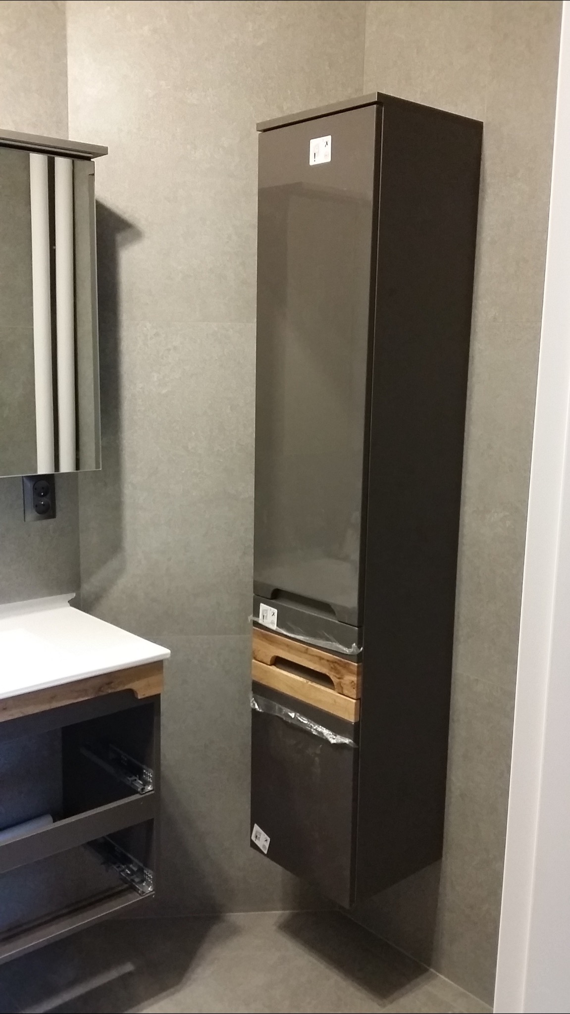 Nowoczesna łazienka z wiszącą szafką w kolorze grafitowym, z drewnianymi szufladami w folii ochronnej, obok umywalki z szafką w podobnym stylu, szara ściana z płytek.