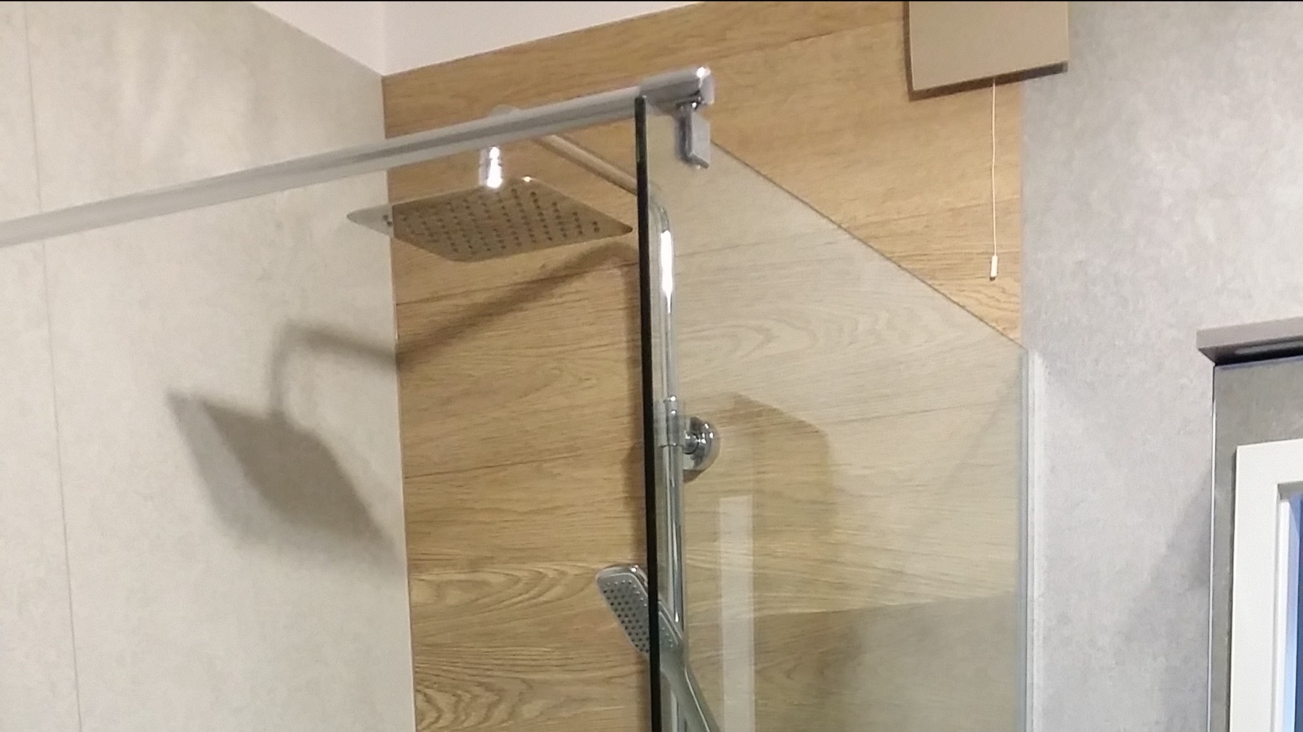 Szklana kabina prysznicowa z deszczownicą i słuchawką, wykończona drewnopodobną ścianką i szarą płytką, widoczny cień deszczownicy na ścianie.