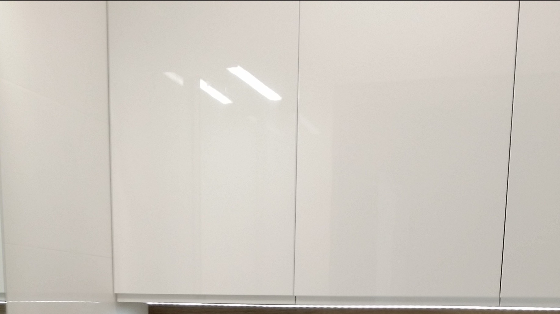 Białe, lakierowane fronty szafek kuchennych z widocznymi odbiciami światła, dolna krawędź szafek podświetlona listwą LED.