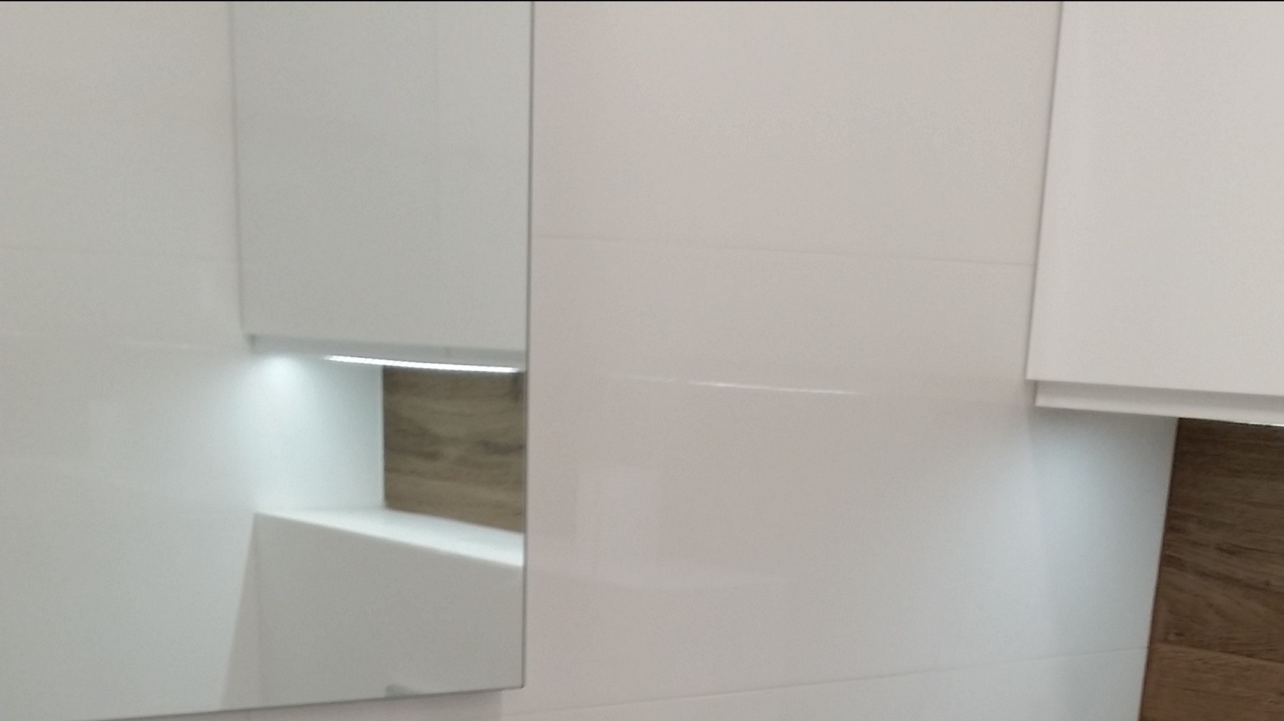 Biała szafka łazienkowa z podświetleniem LED i drewnopodobnym panelem w tle, odbicie w lustrze.