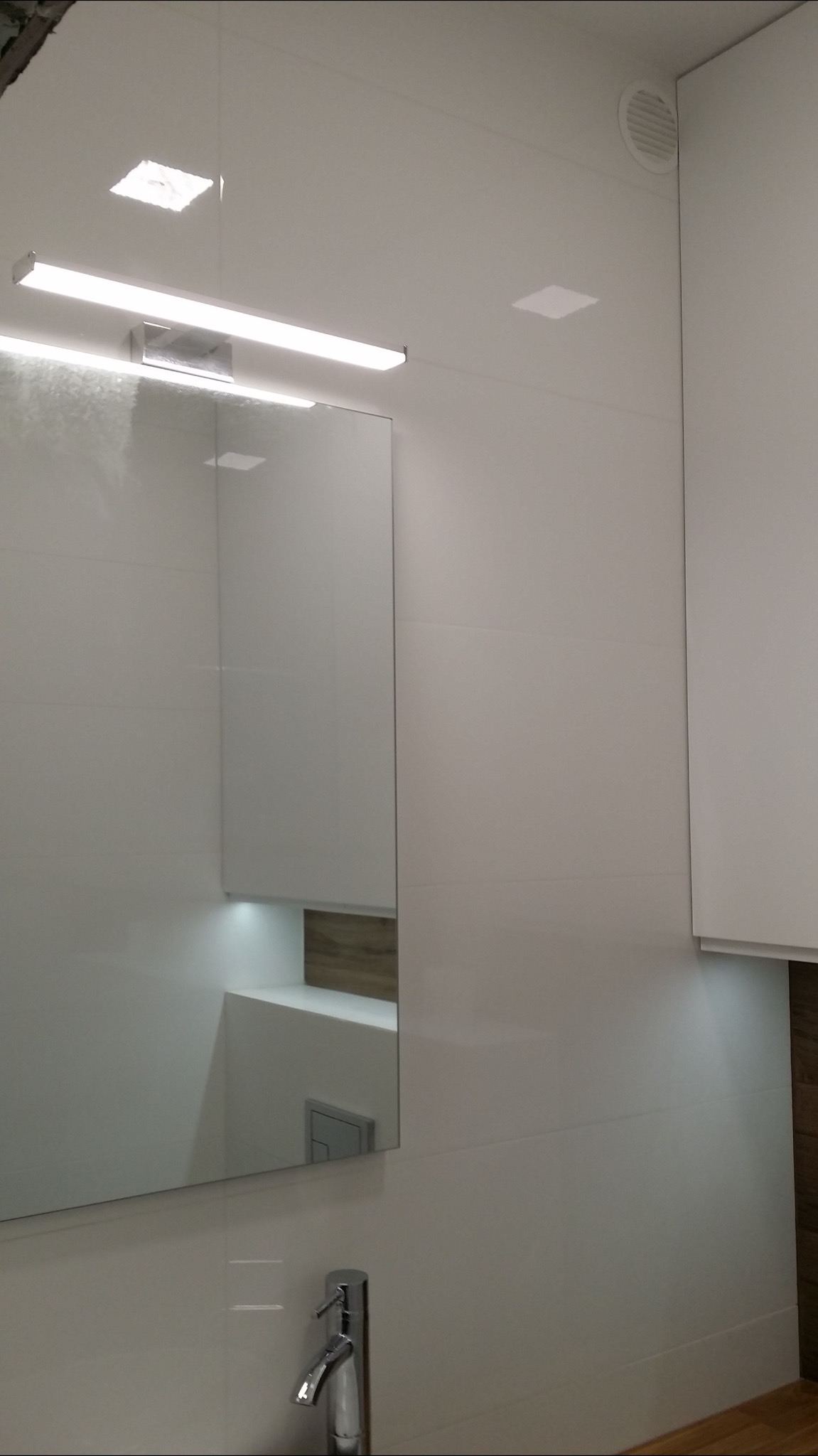 Nowoczesna łazienka z białymi, błyszczącymi płytkami, podświetlanym lustrem i minimalistyczną armaturą. Widoczny fragment szafki z drewnianym akcentem i wentylacja sufitowa.