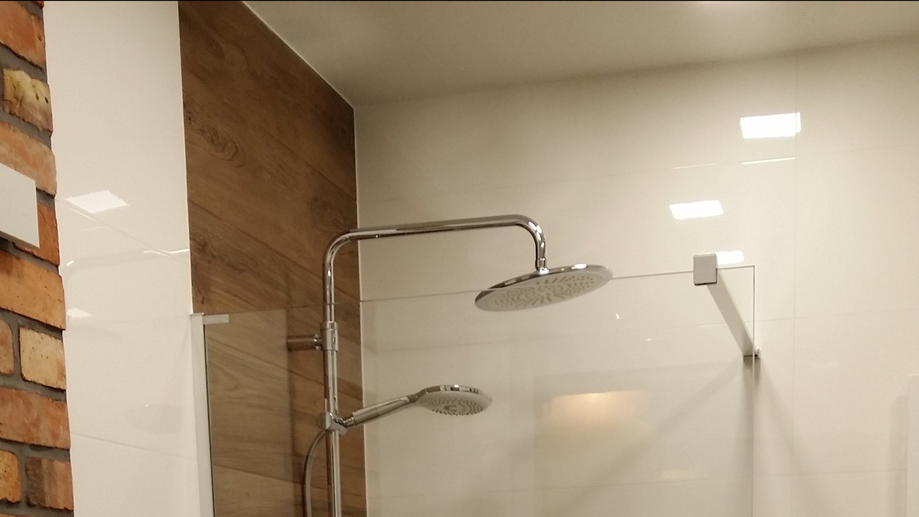 Nowoczesna kabina prysznicowa z deszczownicą i panelem z imitacji drewna, kontrastująca z fragmentem ściany wykończonej czerwoną cegłą.