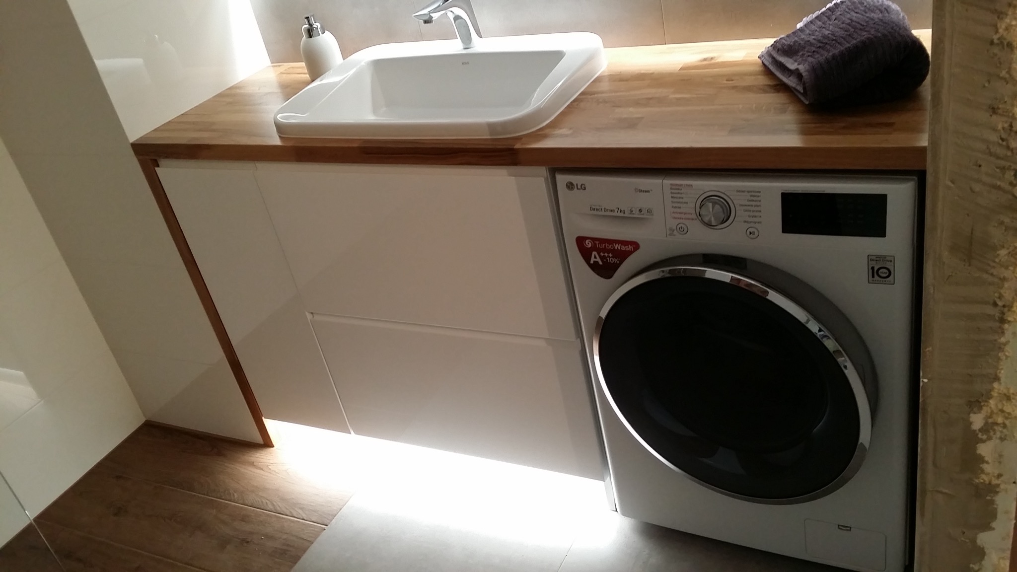 Nowoczesna łazienka z białą umywalką nablatową na drewnianym blacie, pod którym znajduje się szafka z połyskiem i pralka LG, całość oświetlona ciepłym światłem.