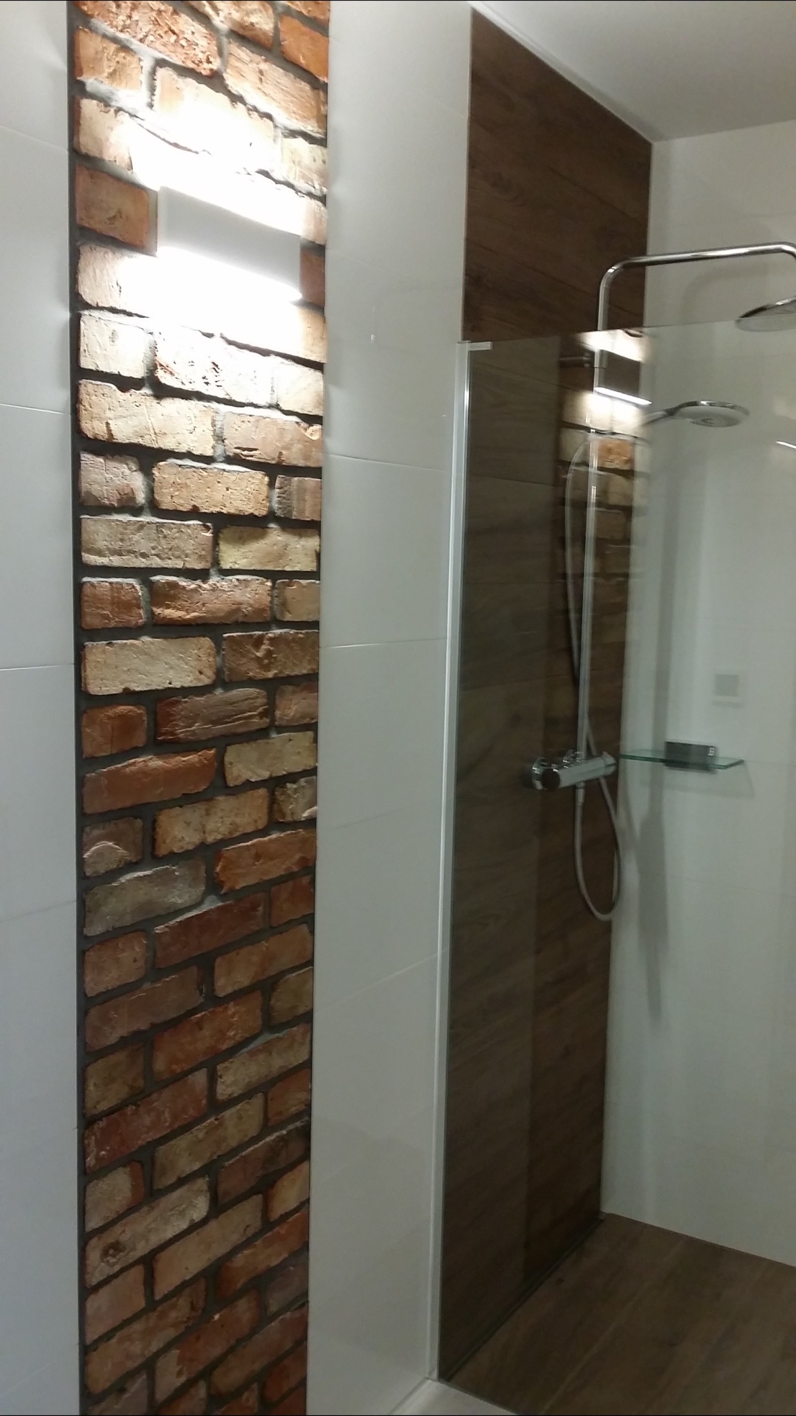 Nowoczesna łazienka z kabiną prysznicową, wykończona białymi płytkami, panelem z imitacją drewna i ścianą z dekoracyjnej cegły z podświetleniem.