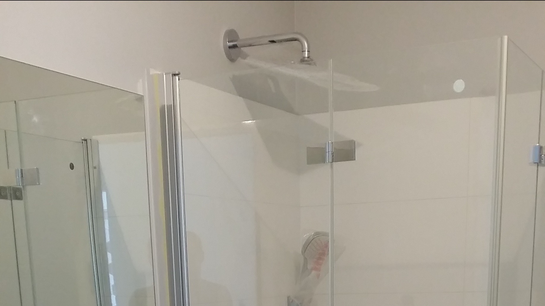 Nowoczesna kabina prysznicowa z chromowaną armaturą i szklanymi drzwiami, widoczne zawiasy i deszczownica.