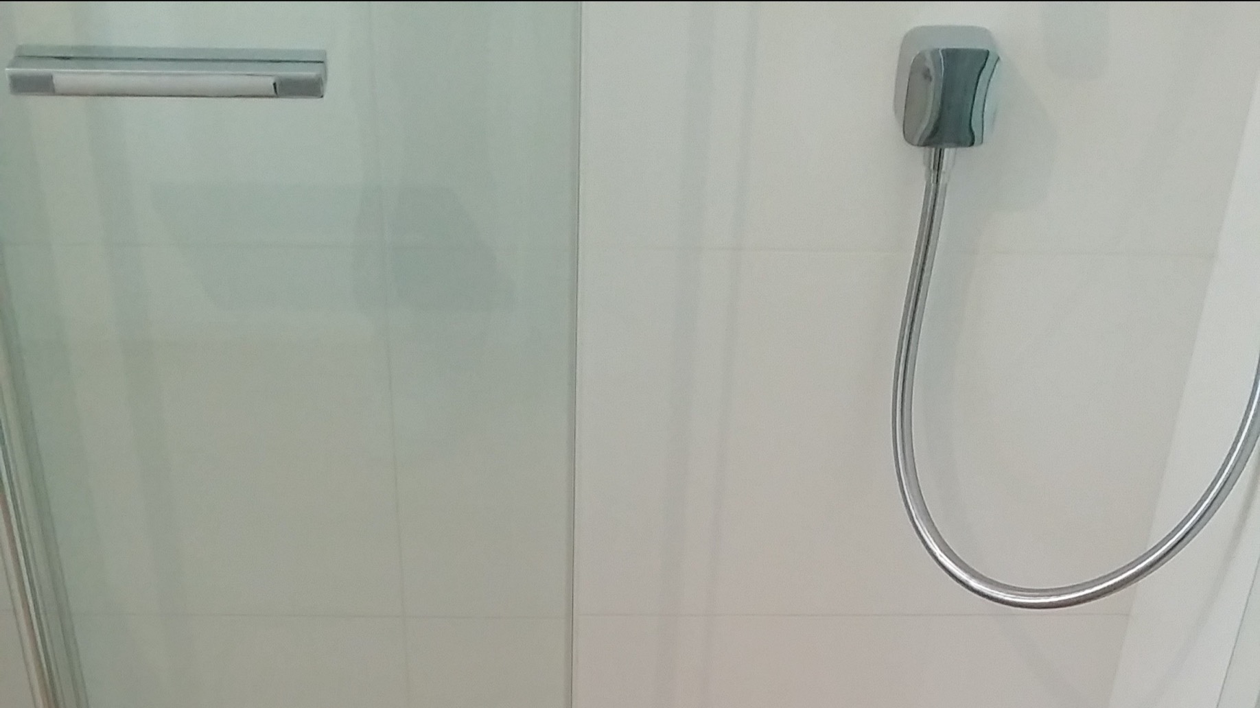 Szczegółowe ujęcie kabiny prysznicowej z chromowanym uchwytem i słuchawką prysznicową na tle białych, kwadratowych płytek.