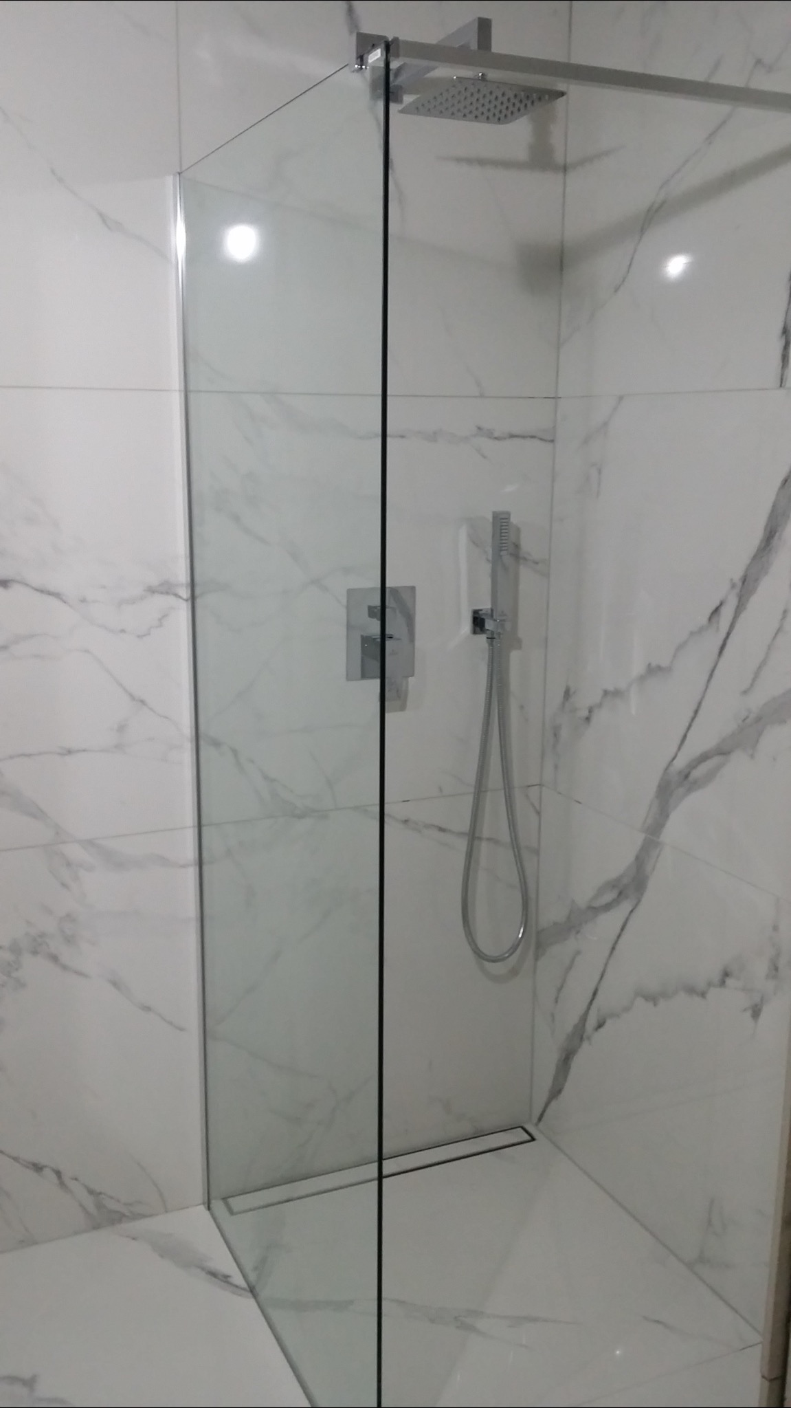 Kabina prysznicowa typu walk-in z chromowaną armaturą i odpływem liniowym, wykończona białymi płytkami imitującymi marmur Carrara.