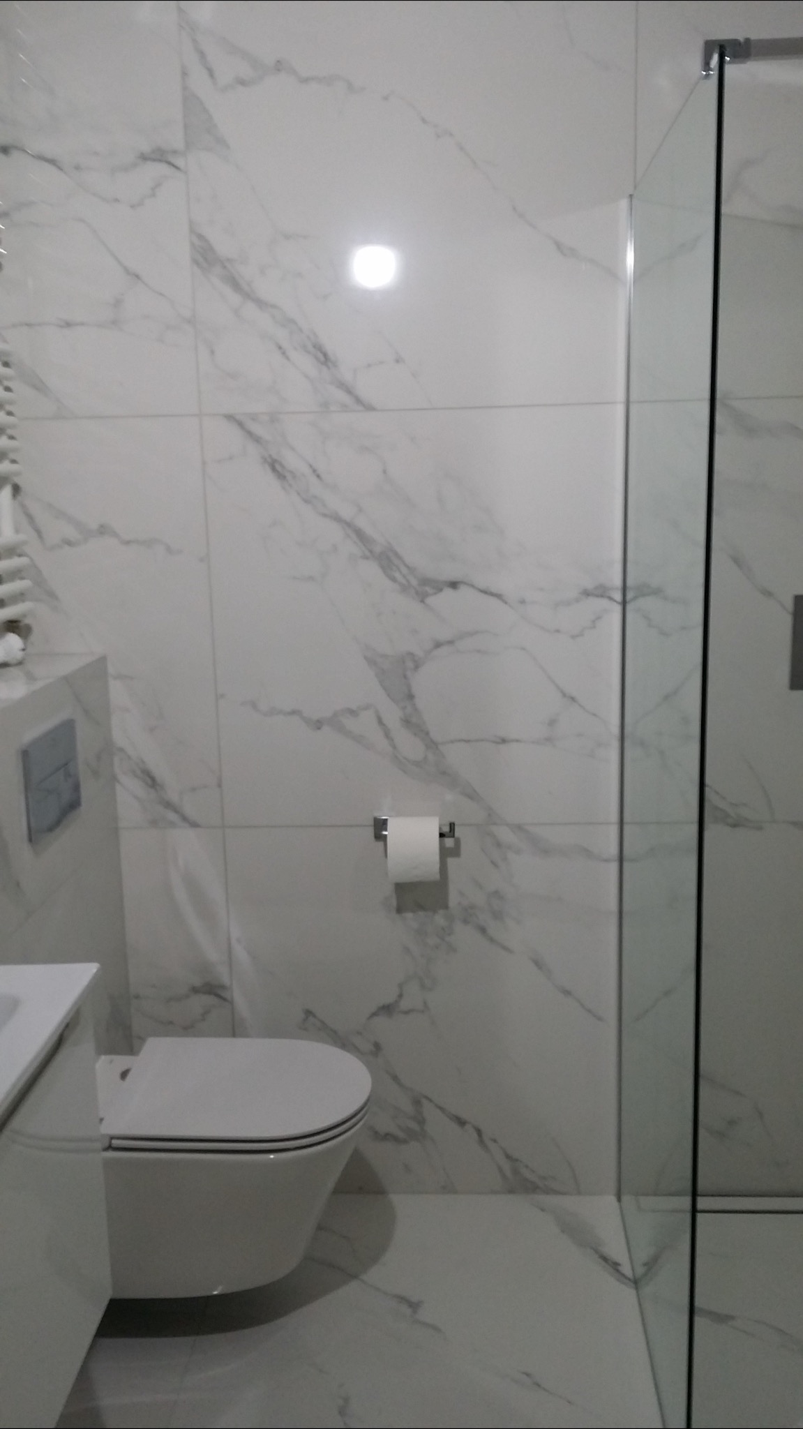 Nowoczesna łazienka z białymi płytkami imitującymi marmur, podwieszana toaleta, szklana kabina prysznicowa, uchwyt na papier toaletowy z rolką.