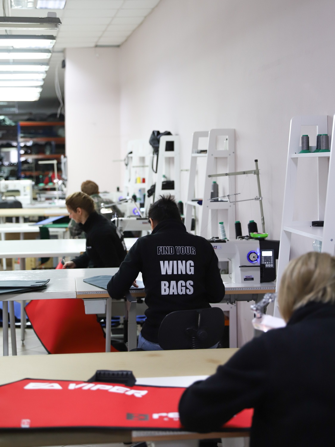 Pracownia krawiecka: Szwaczka przy maszynie, szyje pokrowce, na plecach bluzy napis 'FIND YOUR WING BAGS', widoczne półki z materiałami i nićmi.