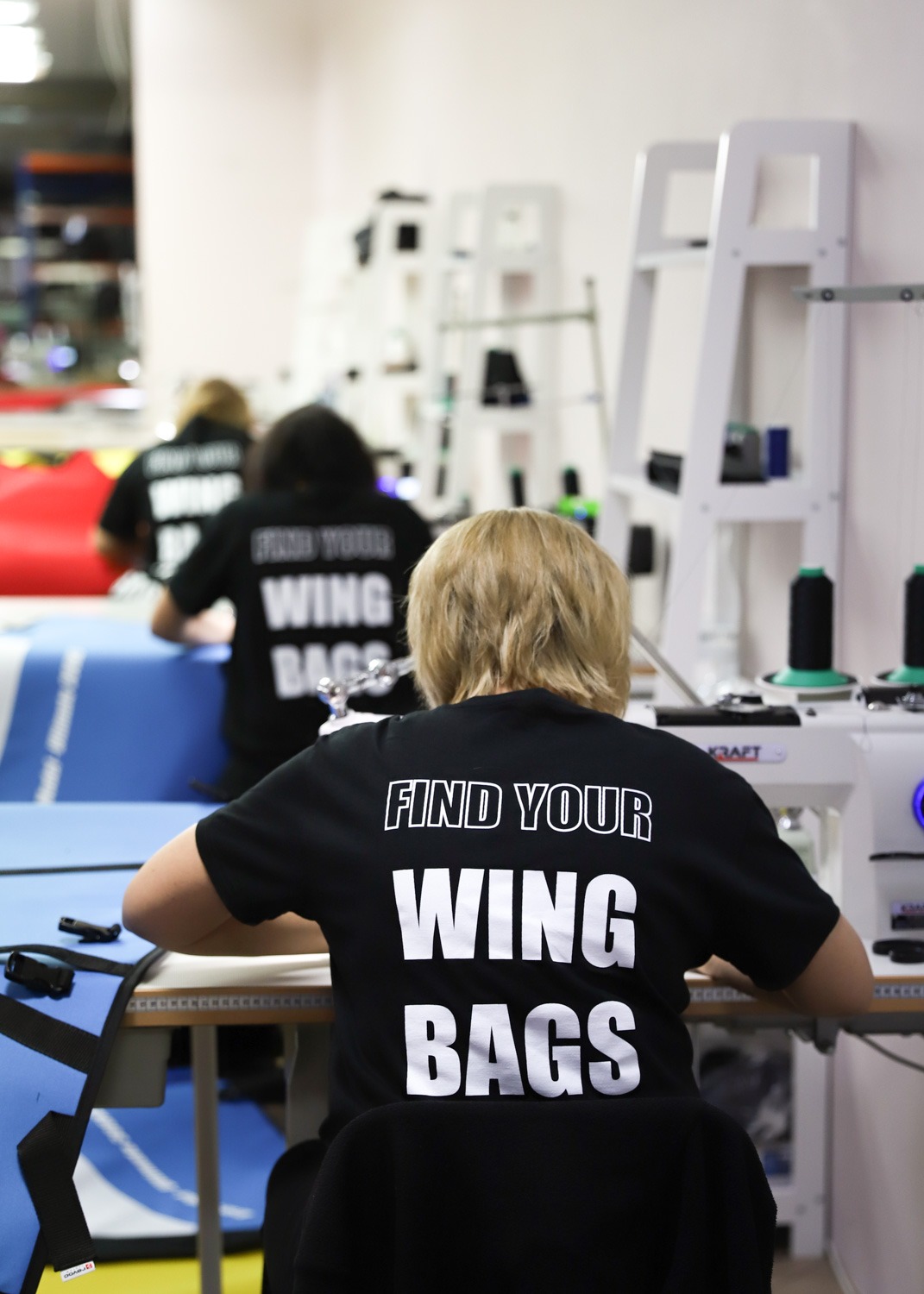 Pracownice szwalni w czarnych koszulkach z napisem 'FIND YOUR WING BAGS' siedzą przy maszynach do szycia, w tle regały z akcesoriami i materiałami.