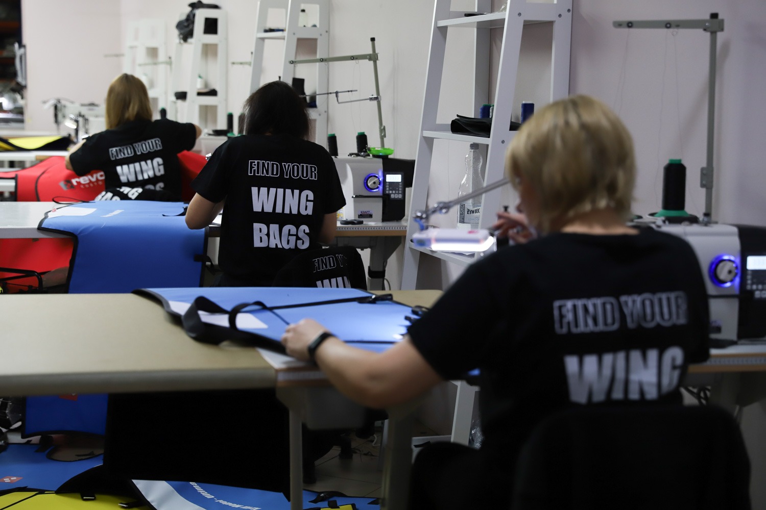 Pracownice szwalni przy stanowiskach z maszynami do szycia, szyjące niebieskie pokrowce z czarną lamówką. Na plecach mają czarne koszulki z napisem 'FIND YOUR WING BAGS'.