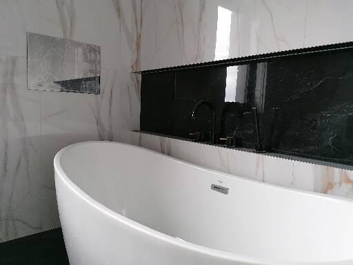 Nowoczesna łazienka z wolnostojącą białą wanną owalną, czarną armaturą i marmurowymi płytkami na ścianach z ukrytym oświetleniem.