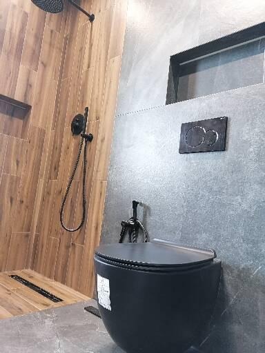 Nowoczesna łazienka z czarną miską WC, prysznicem z deszczownicą i drewnopodobnymi płytkami na ścianie, szare płytki imitujące beton, czarna armatura.