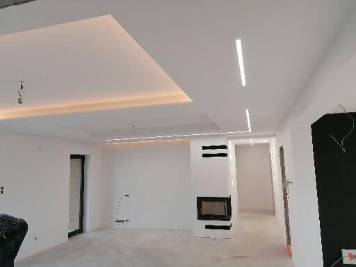 Wnętrze w trakcie prac wykończeniowych, widoczne białe ściany, sufit z oświetleniem LED, kominek z czarnymi elementami dekoracyjnymi, oraz przygotowane podłączenia elektryczne na ścianach.