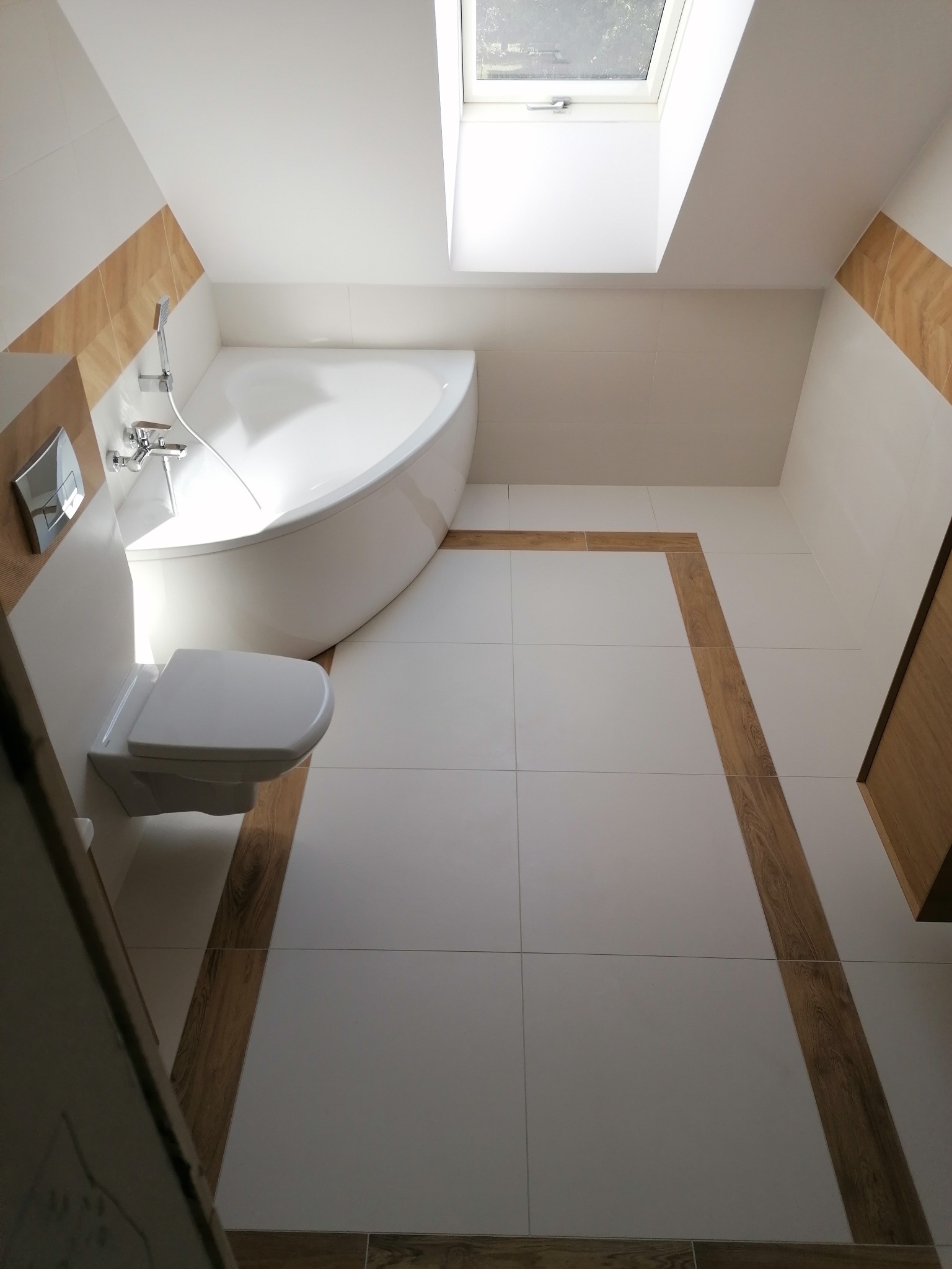 Nowoczesna łazienka na poddaszu z oknem dachowym, białe płytki z drewnopodobnym akcentem, narożna wanna i podwieszana toaleta.