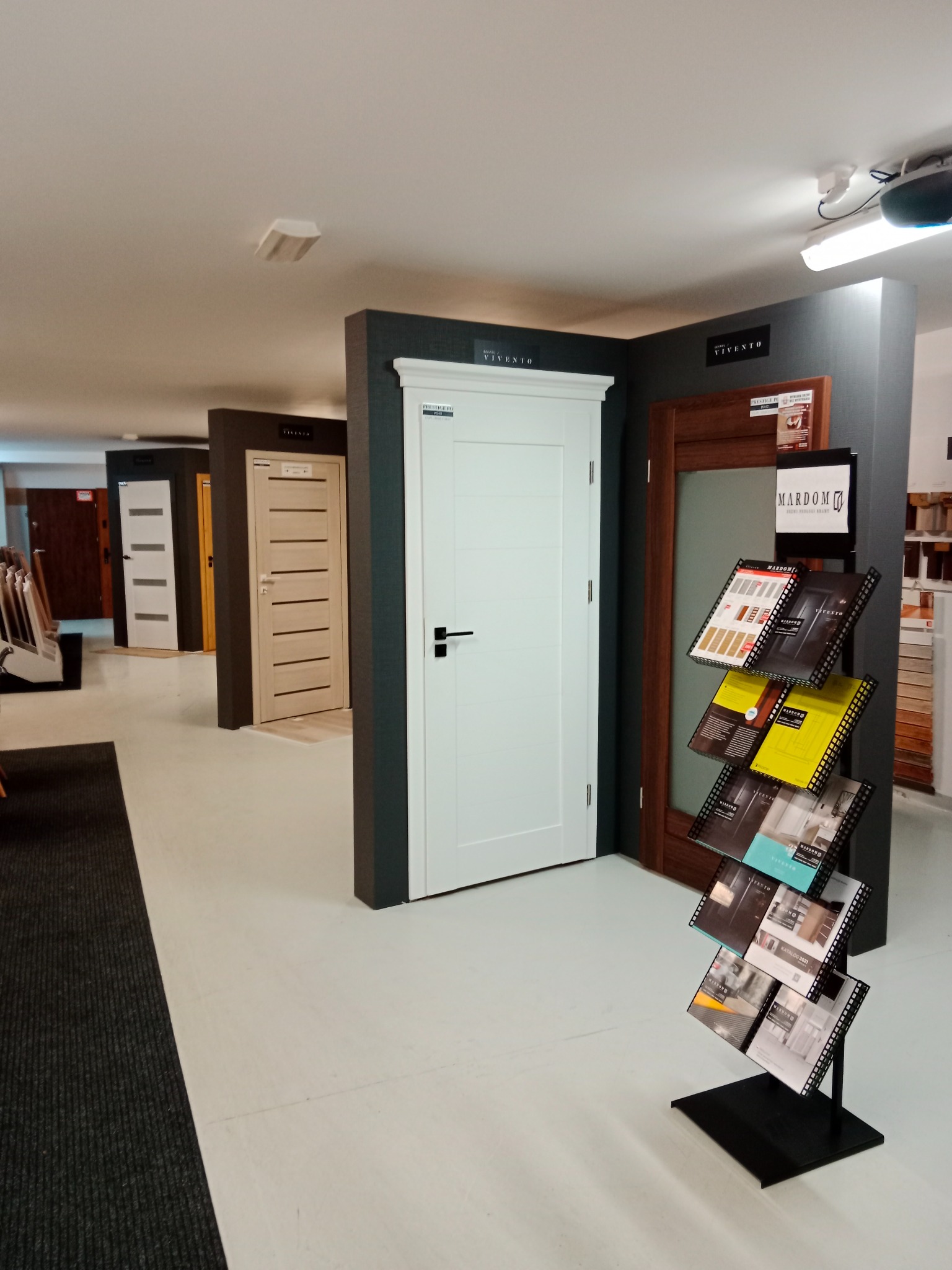 Ekspozycja modeli drzwi wewnętrznych w salonie, widoczne różne wzory i kolory, w tym białe drzwi z czarną klamką na pierwszym planie, oraz stojak z katalogami producenta.