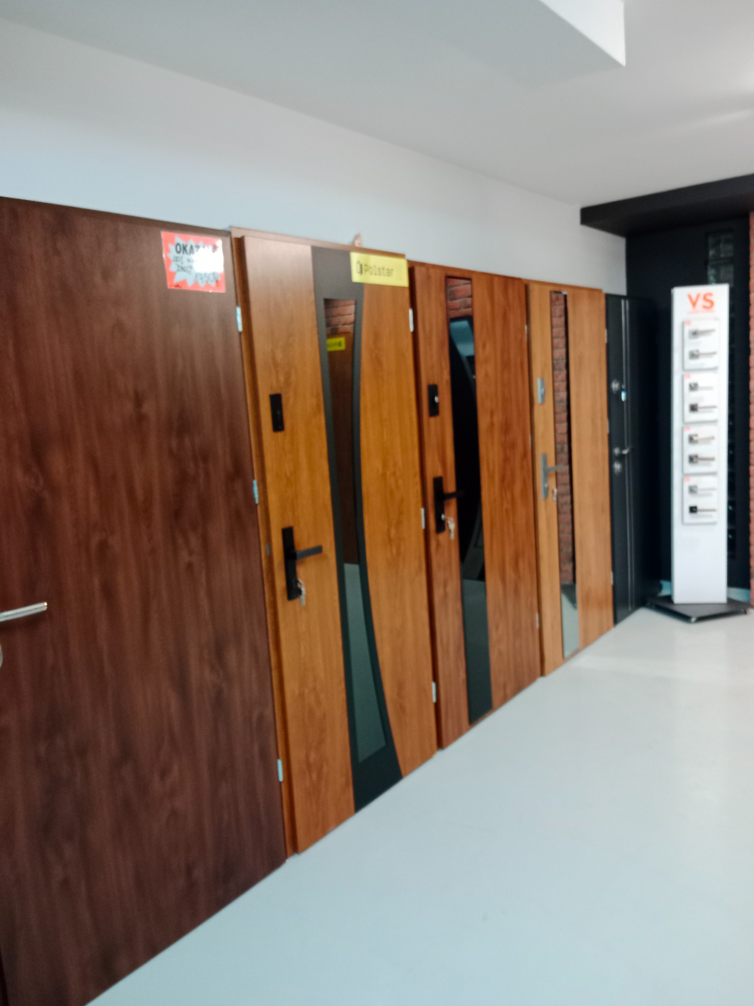 Ekspozycja kilku modeli drzwi wewnętrznych i zewnętrznych w salonie sprzedaży, widoczne różne wykończenia i kolory, w tle tablica informacyjna VS.