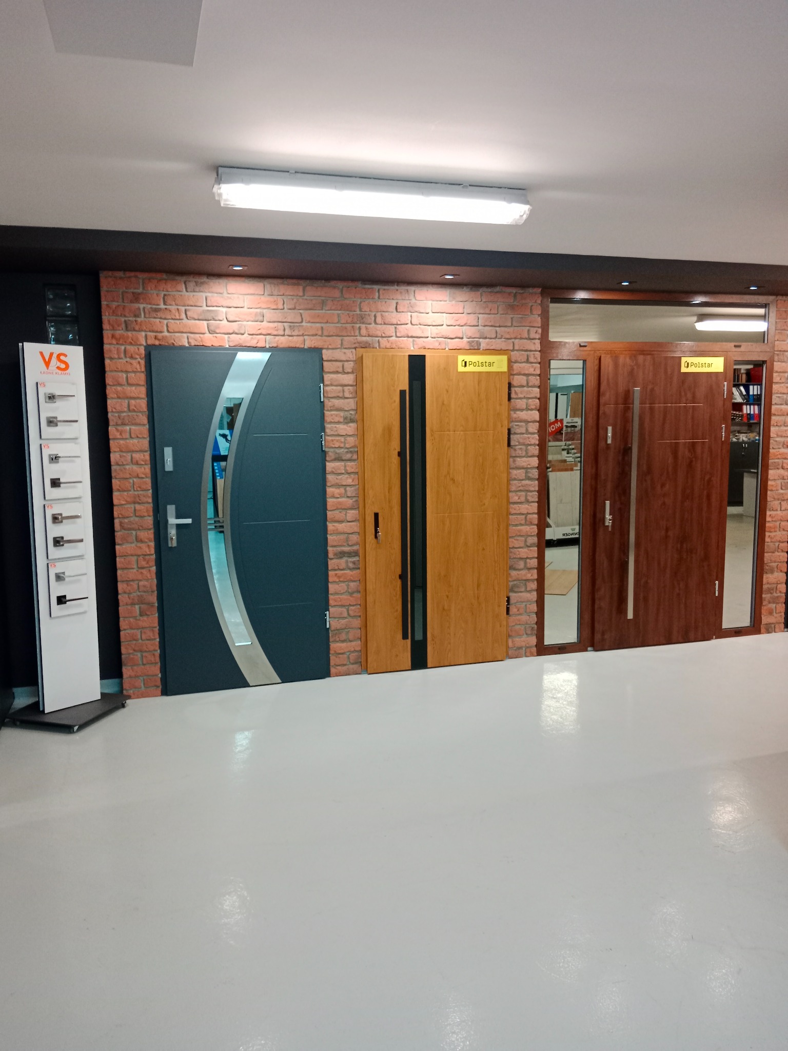 Ekspozycja trzech różnych modeli drzwi z szybami i bez, na tle ściany z cegły w salonie sprzedaży. Po lewej ekspozytor z klamkami.