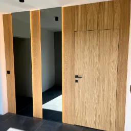 Nowoczesne wykończenie wnętrza: drewniana boazeria z ukrytymi drzwiami i podświetleniem LED, czarne lampy sufitowe i ciemna, matowa podłoga.