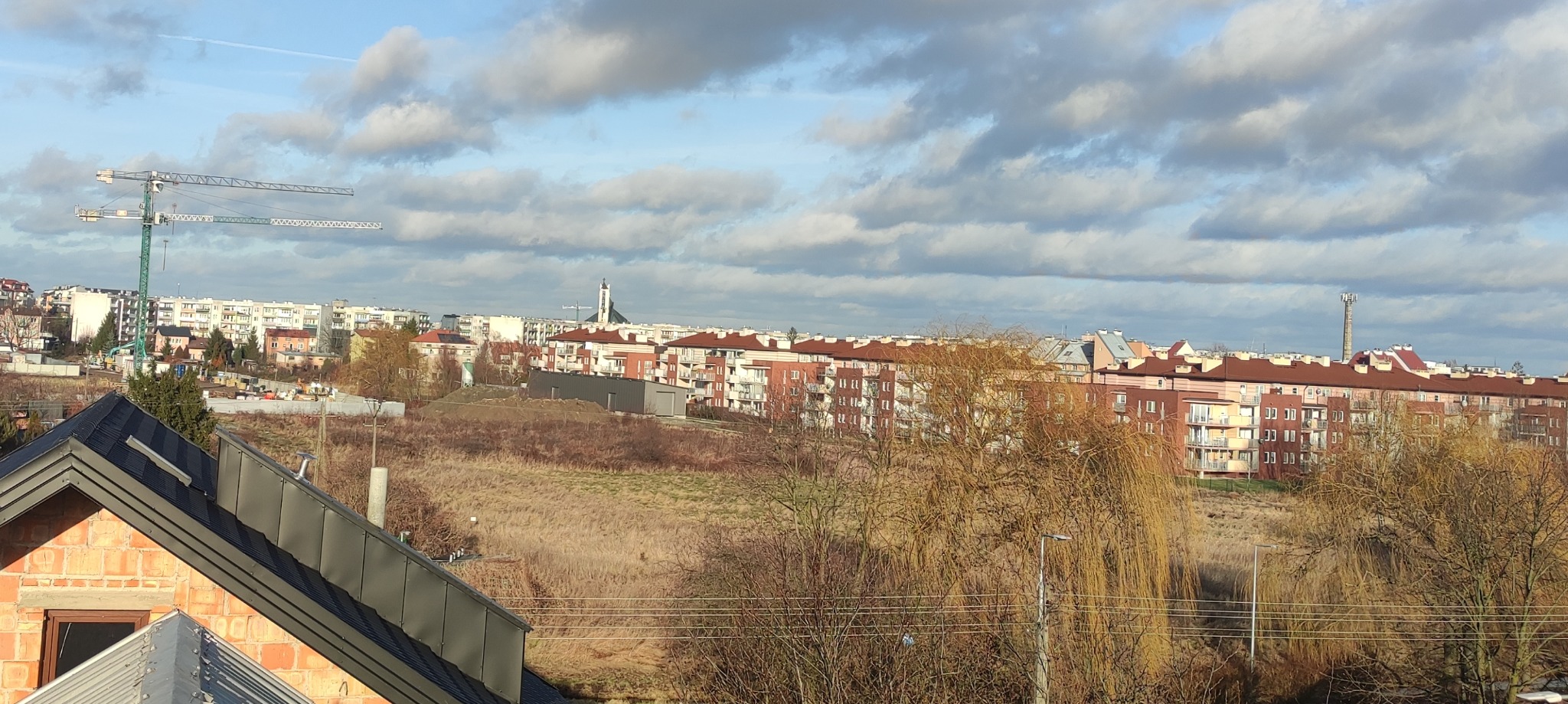 Widok na osiedle mieszkaniowe z nowym dachem z blachy trapezowej na pierwszym planie, w tle budowa z żurawiem i kościół.