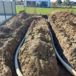 Wykop liniowy w ziemi z ułożonymi rurami, widok z góry na tle ogrodzenia i budynku mieszkalnego w słoneczny dzień.