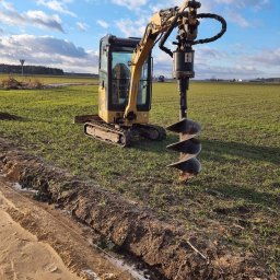 ASTRAKOP - Żółta mini koparka z wiertłem ziemnym w trakcie pracy na polu, obok wykopany rów z widoczną linką wyznaczającą linię, błękitne niebo z chmurami w tle.
