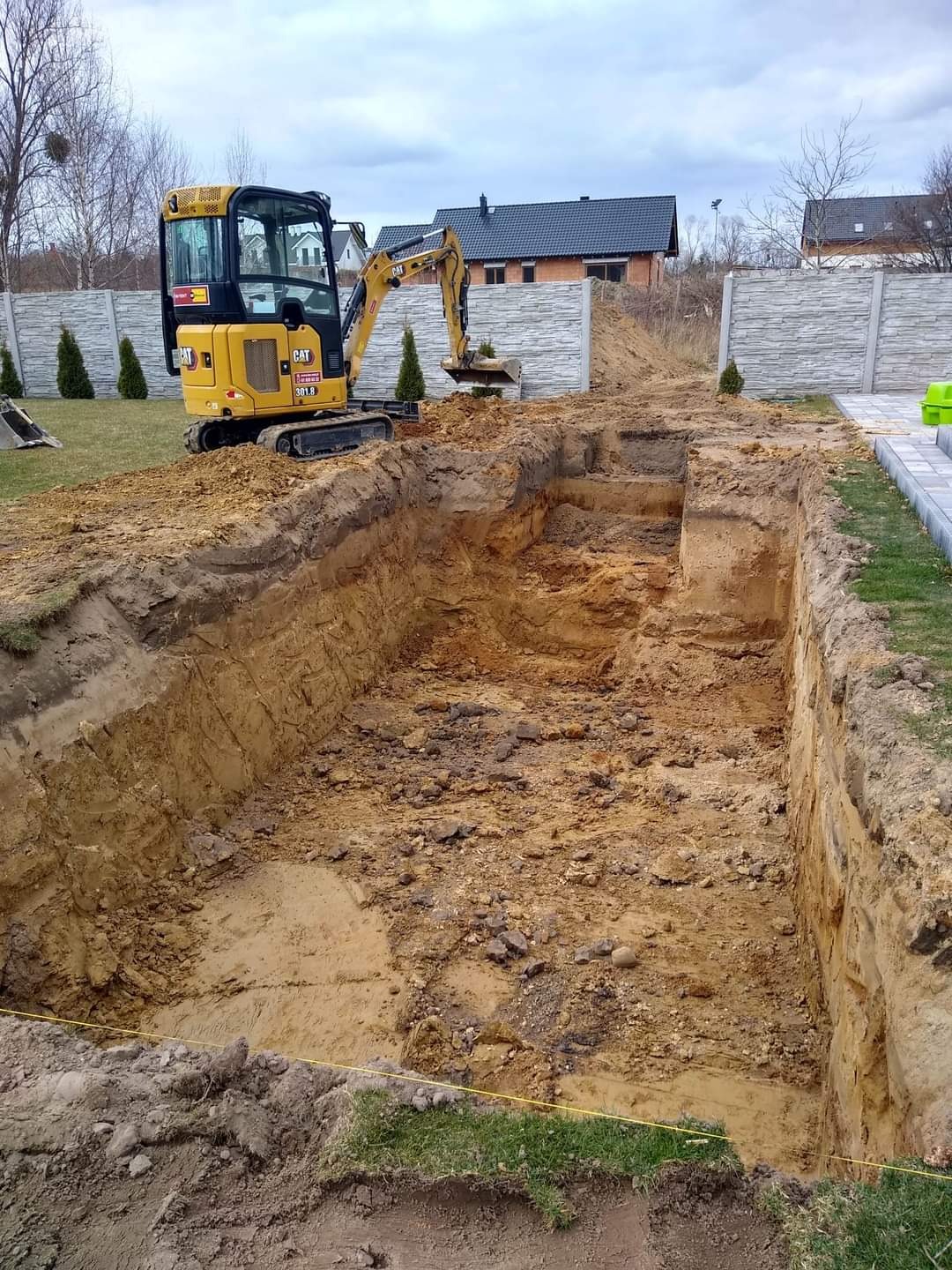 Żółta mini koparka CAT 301.8 przy głębokim wykopie w ziemi, widoczne ślady po pracy łyżką, w tle betonowe ogrodzenie i dom.